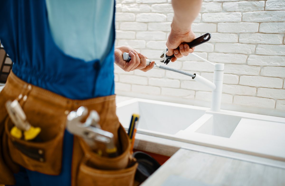 Male plumber in uniform fixing faucet in the kitchen. Handyman with toolbag repair sink, sanitary equipment service at home